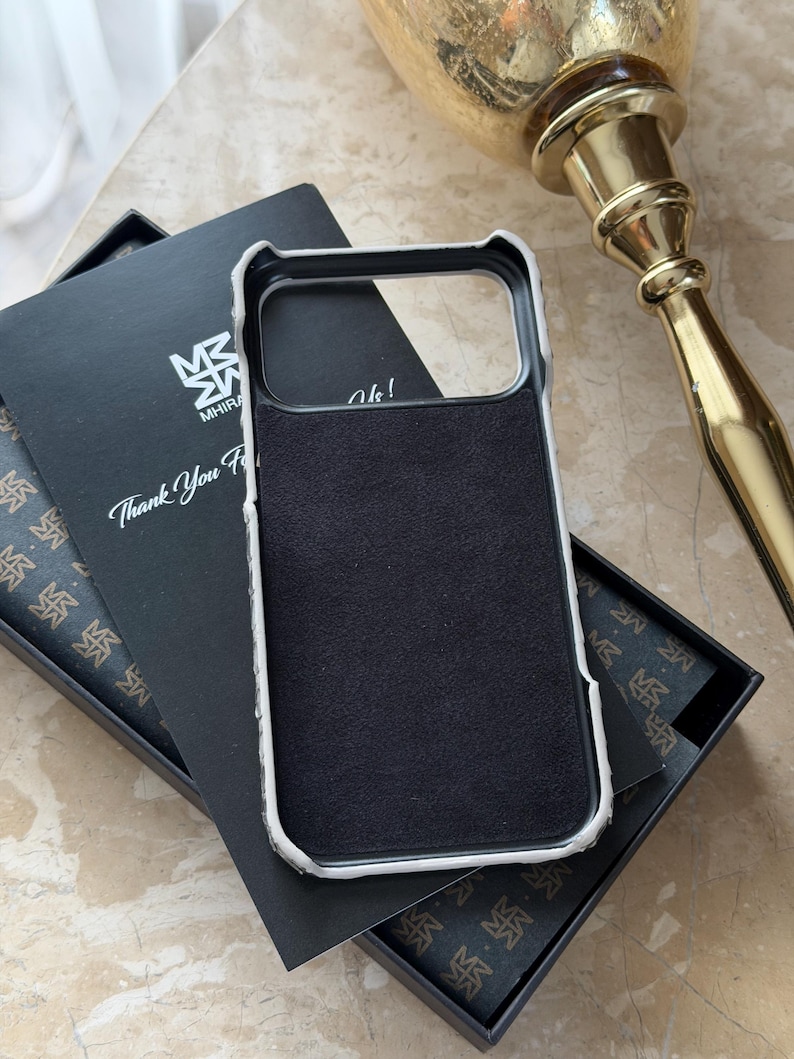 May include: A black phone case with a white border, resting in an open black box with a patterned interior. The case has a black suede-like interior. A card with the text "Thank You For" is visible.