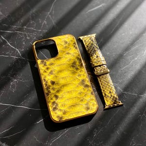 Pu&ograve; includere: Una custodia per telefono gialla e marrone con texture a pelle di serpente con una cornice color oro e un cinturino per orologio abbinato. La custodia e il cinturino sono su una superficie scura e marmorizzata.