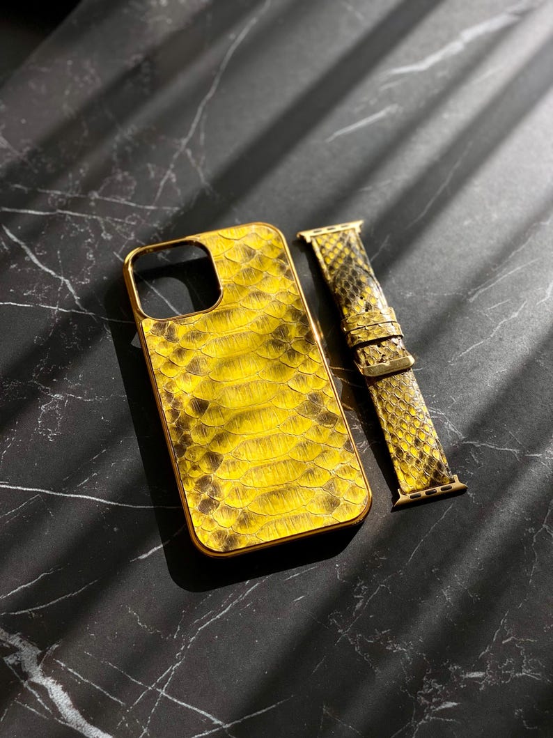 Pu&ograve; includere: Una custodia per telefono gialla e marrone con texture a pelle di serpente con una cornice color oro e un cinturino per orologio abbinato. La custodia e il cinturino sono su una superficie scura e marmorizzata.