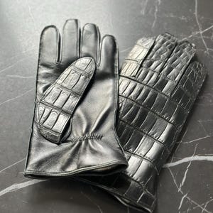 May include: A pair of black leather gloves with a crocodile pattern. The gloves are folded together and the crocodile pattern is visible on both the back and the palm of the gloves.