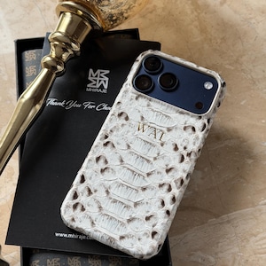 May include: White and brown snakeskin-patterned phone case with the initials "WAT" on the front. The case is on top of a black box with the text "Thank You For Choosing" and the brand name "MHIRAJE". A gold-colored decorative object is in the background.
