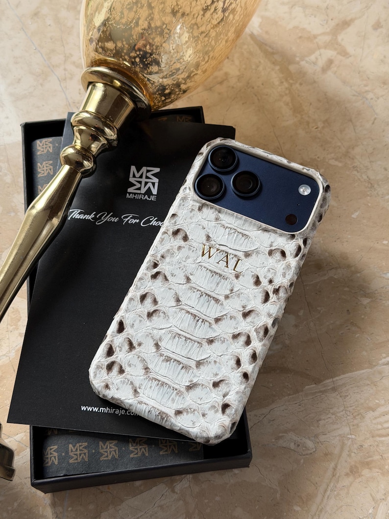 May include: White and brown snakeskin-patterned phone case with the initials "WAT" on the front. The case is on top of a black box with the text "Thank You For Choosing" and the brand name "MHIRAJE". A gold-colored decorative object is in the background.
