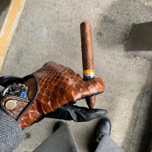 May include: A person wearing brown leather gloves with a black leather cuff holds a brown cigar with a yellow band. The cigar is held between the thumb and forefinger of the right hand. The person is wearing a gray suit jacket and black shoes.