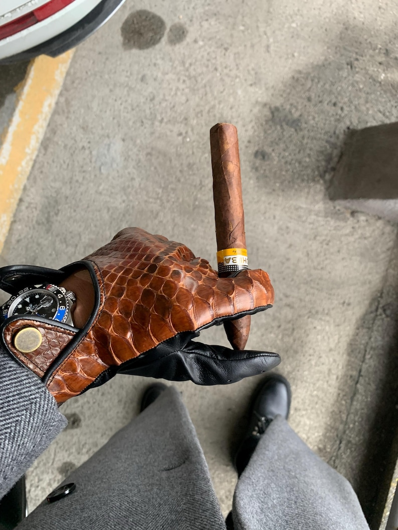 May include: A person wearing brown leather gloves with a black leather cuff holds a brown cigar with a yellow band. The cigar is held between the thumb and forefinger of the right hand. The person is wearing a gray suit jacket and black shoes.