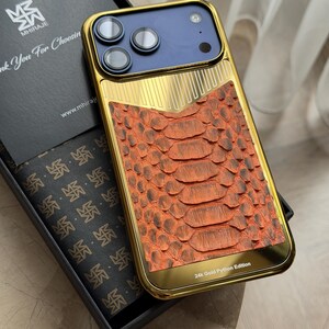 Op de afbeelding: Een goudkleurige telefoonhoes met een textuurontwerp van oranje pythonskin. De hoes heeft een blauw cameragedeelte en wordt gepresenteerd in een zwarte doos. De tekst "24k Gold Python Edition" is zichtbaar.