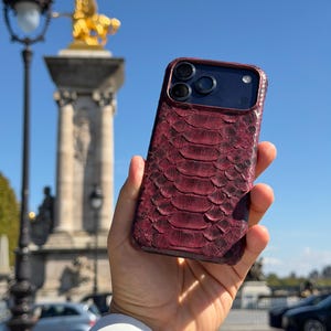May include: A smartphone case featuring a deep burgundy, textured snakeskin pattern. The case has a dark camera cutout area with three circular lenses and a flash. The surface of the case displays a distinct, overlapping scale design.