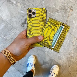 Pu&ograve; includere: Una custodia per telefono e un portafoglio abbinato con motivo a pelle di serpente gialla sono tenuti in mano. Il portafoglio contiene una carta di credito. Un orologio d'oro &egrave; al polso. Sono visibili scarpe da ginnastica bianche con dettagli blu.