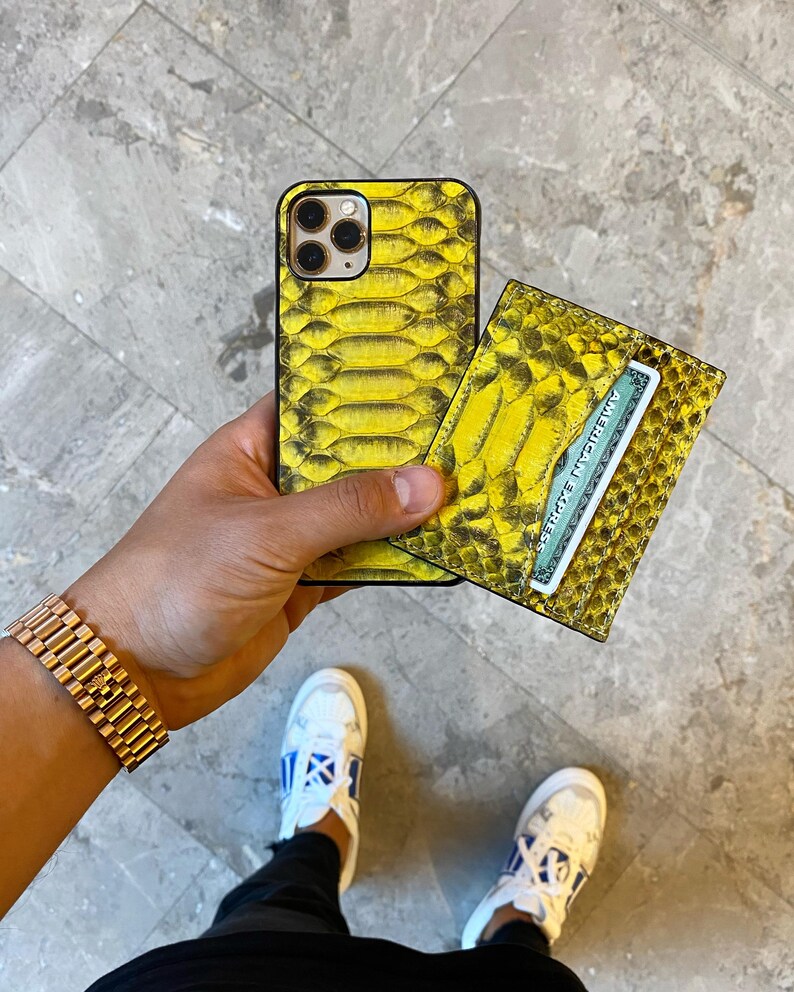 Pu&ograve; includere: Una custodia per telefono e un portafoglio abbinato con motivo a pelle di serpente gialla sono tenuti in mano. Il portafoglio contiene una carta di credito. Un orologio d'oro &egrave; al polso. Sono visibili scarpe da ginnastica bianche con dettagli blu.