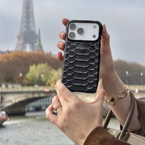 Pode incluir: Uma capa de smartphone preta com um padr&atilde;o texturizado de escamas sobrepostas cobrindo a parte traseira. A capa apresenta um recorte retangular para o m&oacute;dulo da c&acirc;mera, que cont&eacute;m tr&ecirc;s lentes circulares e um flash, revelando o telefone branco.