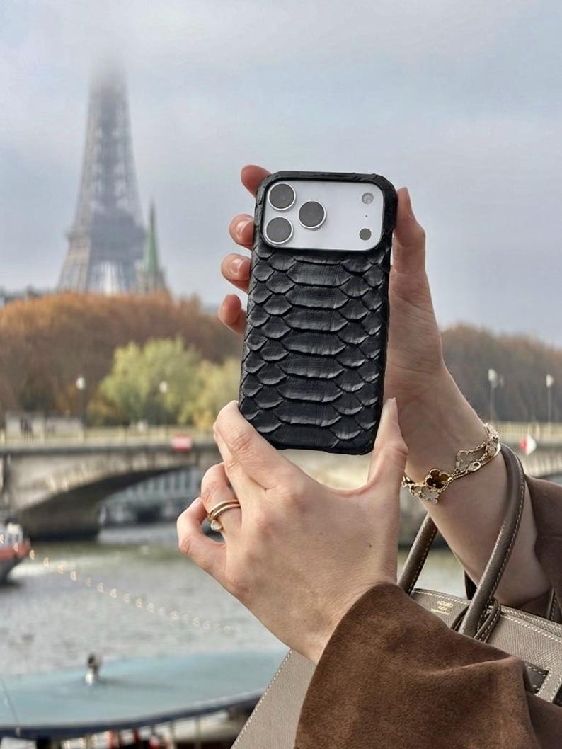 Pode incluir: Uma capa de smartphone preta com um padr&atilde;o texturizado de escamas sobrepostas cobrindo a parte traseira. A capa apresenta um recorte retangular para o m&oacute;dulo da c&acirc;mera, que cont&eacute;m tr&ecirc;s lentes circulares e um flash, revelando o telefone branco.