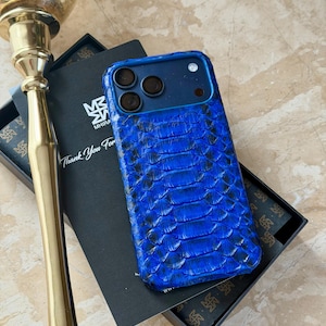 May include: A blue phone case with a snakeskin pattern. The case has a dark blue frame and three camera cutouts. The case is in a black box with the text "Thank You For" and a logo. A gold candlestick is in the background.