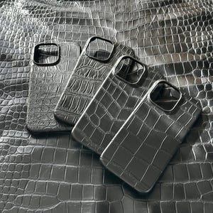 Op de afbeelding: Vier zwarte smartphonehoesjes met een gestructureerd krokodillenleerpatroon. Elk hoesje heeft een rechthoekige uitsparing voor de cameralens in de linkerbovenhoek. De hoesjes zijn in een verspringende, overlappende formatie geplaatst.