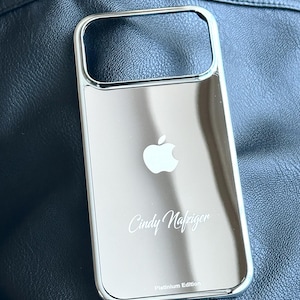 Puede incluir: Funda de tel&eacute;fono plateada con superficie reflectante. Presenta un logotipo de Apple blanco y el nombre "Cindy Nafziger" en cursiva. El texto "Platinum Edition" est&aacute; en la parte inferior.