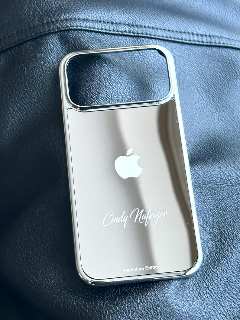 Puede incluir: Funda de tel&eacute;fono plateada con superficie reflectante. Presenta un logotipo de Apple blanco y el nombre "Cindy Nafziger" en cursiva. El texto "Platinum Edition" est&aacute; en la parte inferior.