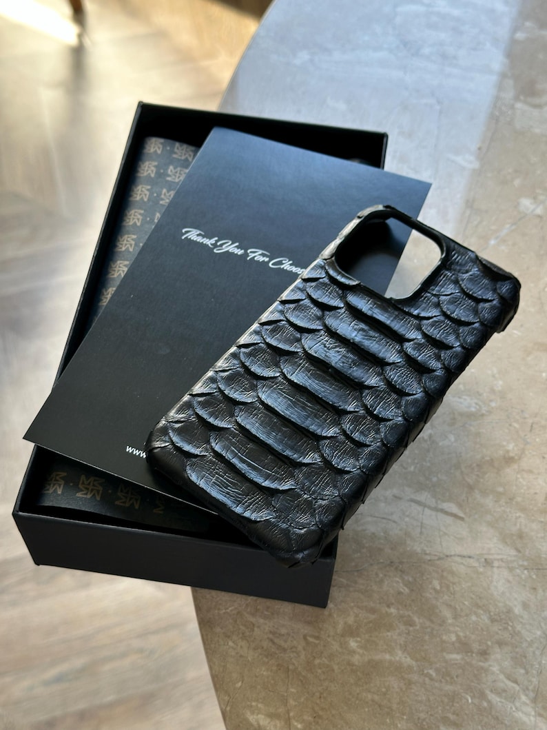May include: Black phone case with a textured, scale-like pattern, resting on a black box with a card that reads "Thank You For Choosing". The case is designed to fit a smartphone and is made of a durable material.