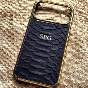 Pode incluir: Uma capa de telefone dourada com um padrão de pele de cobra texturizada azul escuro. As iniciais "SPG" são gravadas em dourado na capa. O fundo é uma superfície dourada com textura de pele de cobra.