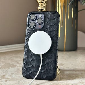 May include: Black textured phone case with a white MagSafe charger attached. The case has a scale-like pattern. The phone case is on a marble surface, with a gold-colored decorative object in the background.