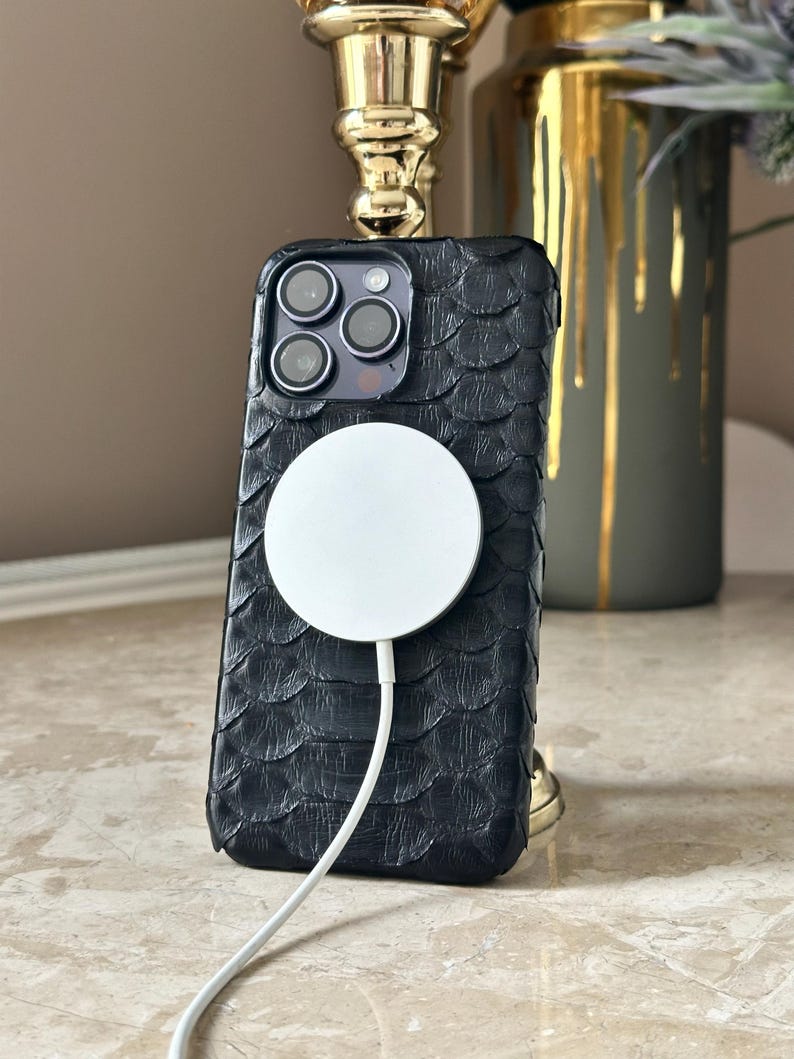 May include: Black textured phone case with a white MagSafe charger attached. The case has a scale-like pattern. The phone case is on a marble surface, with a gold-colored decorative object in the background.