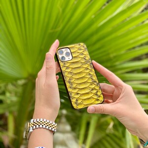 Pu&ograve; includere: Una custodia per telefono gialla e nera con texture a pelle di serpente. La custodia &egrave; tenuta davanti a uno sfondo di foglie verdi. La custodia del telefono ha un ritaglio per la fotocamera e un bordo nero.