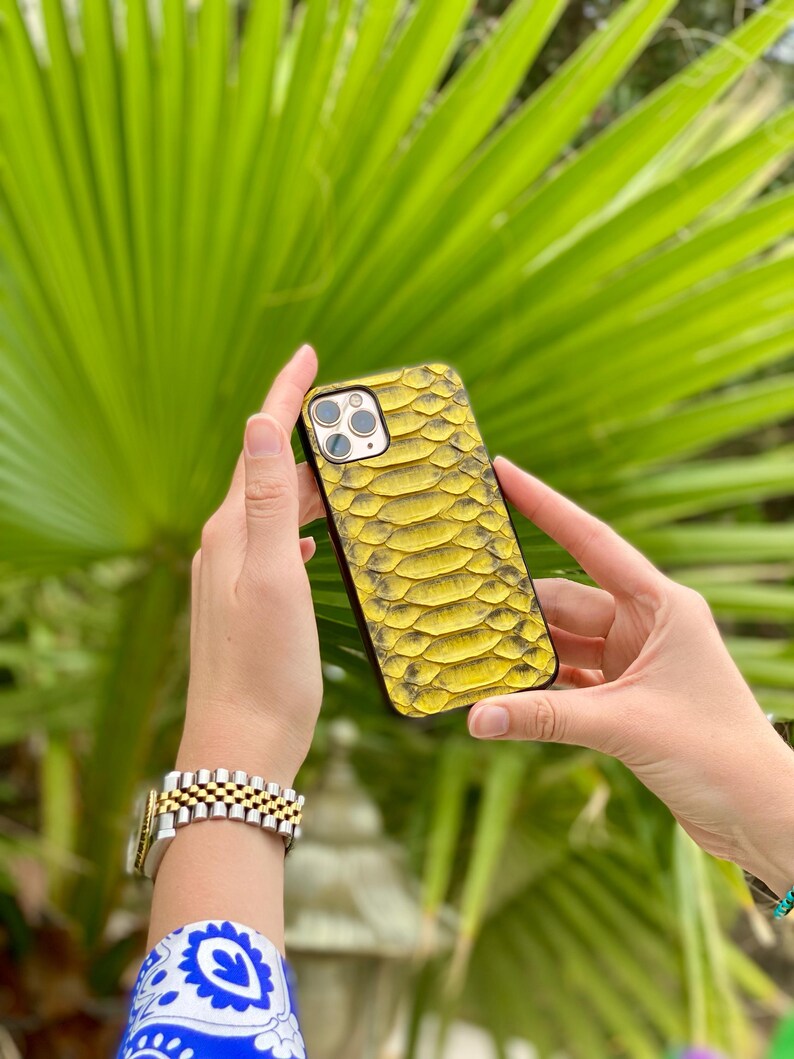 Pu&ograve; includere: Una custodia per telefono gialla e nera con texture a pelle di serpente. La custodia &egrave; tenuta davanti a uno sfondo di foglie verdi. La custodia del telefono ha un ritaglio per la fotocamera e un bordo nero.