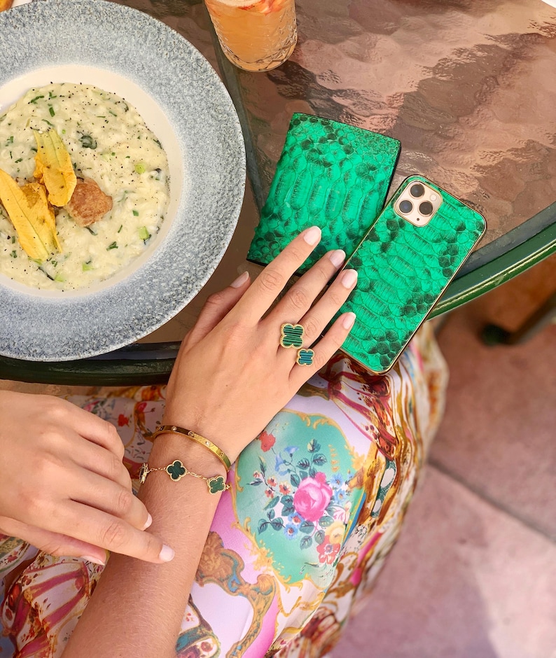 May include: A green snakeskin-textured phone case and matching wallet are displayed on a glass table. A gold bracelet and ring with green clover accents are visible. A plate of risotto and a drink are also on the table.