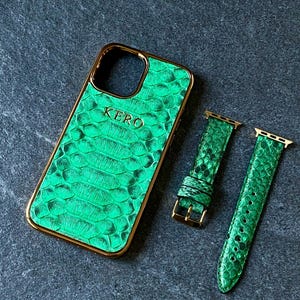 May include: A green snakeskin-textured phone case with gold trim and the name "KERO" embossed on the front. Also included are two green watch bands with gold hardware. The items are displayed on a dark gray surface.