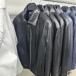 Könnte beinhalten: Eine Reihe schwarzer, blauer und weißer Lederjacken, die an einem Kleiderständer hängen. Die Jacken sind aus Schlangenleder und haben einen Reißverschluss.