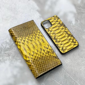 Pu&ograve; includere: Un portafoglio e una custodia per telefono con motivo a pelle di serpente gialla. Il portafoglio &egrave; rettangolare e piegato, mentre la custodia del telefono ha un ritaglio per la fotocamera. Entrambi gli articoli hanno una superficie strutturata e un bordo scuro.