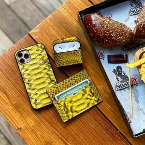 Pu&ograve; includere: Una custodia per telefono, una custodia per AirPods e un porta carte con texture di pelle di serpente gialla sono esposti su una superficie di legno. Il porta carte contiene una carta American Express. La custodia degli AirPods &egrave; aperta.