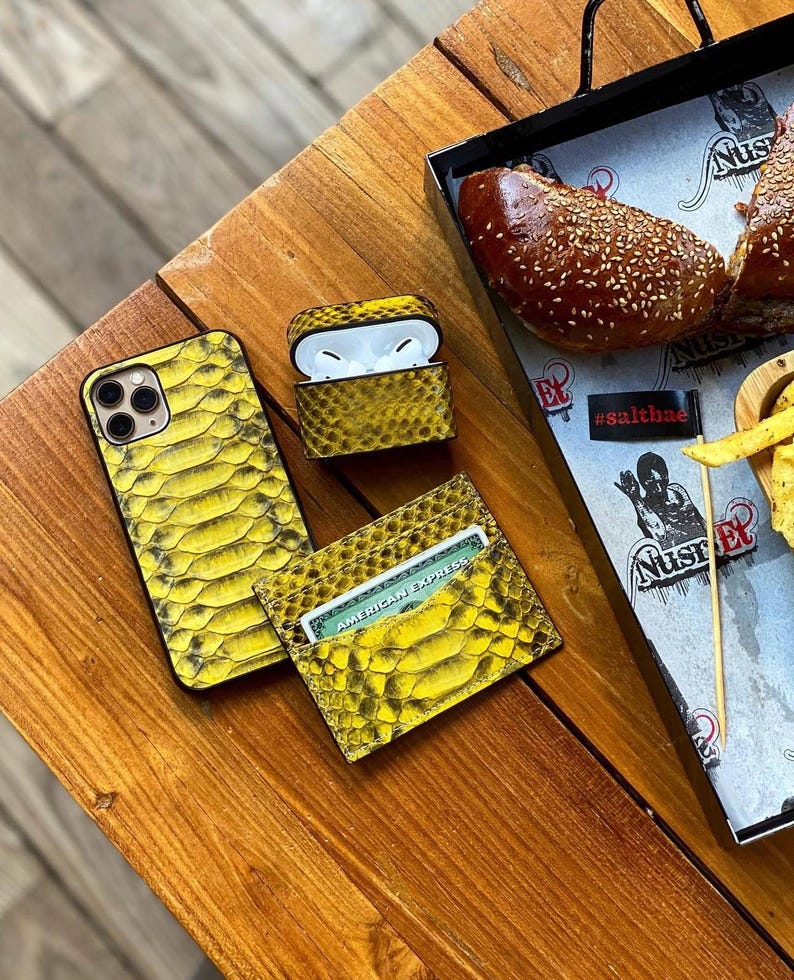 Pu&ograve; includere: Una custodia per telefono, una custodia per AirPods e un porta carte con texture di pelle di serpente gialla sono esposti su una superficie di legno. Il porta carte contiene una carta American Express. La custodia degli AirPods &egrave; aperta.