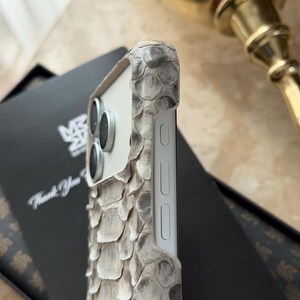 May include: A close-up of a smartphone case with a snakeskin texture in shades of gray and white. The case has a white frame and covers the phone's camera lenses. The case is on top of a black box with the text "Thank You".