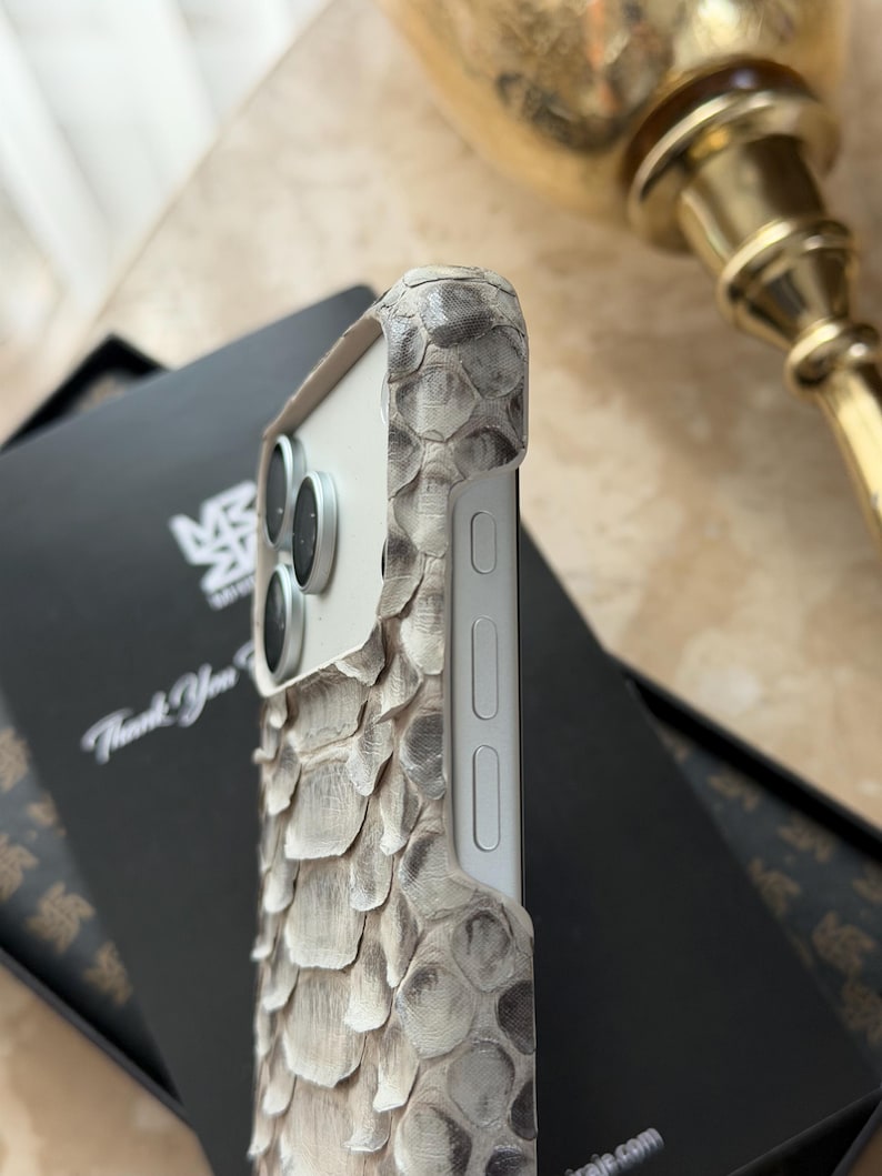 May include: A close-up of a smartphone case with a snakeskin texture in shades of gray and white. The case has a white frame and covers the phone's camera lenses. The case is on top of a black box with the text "Thank You".