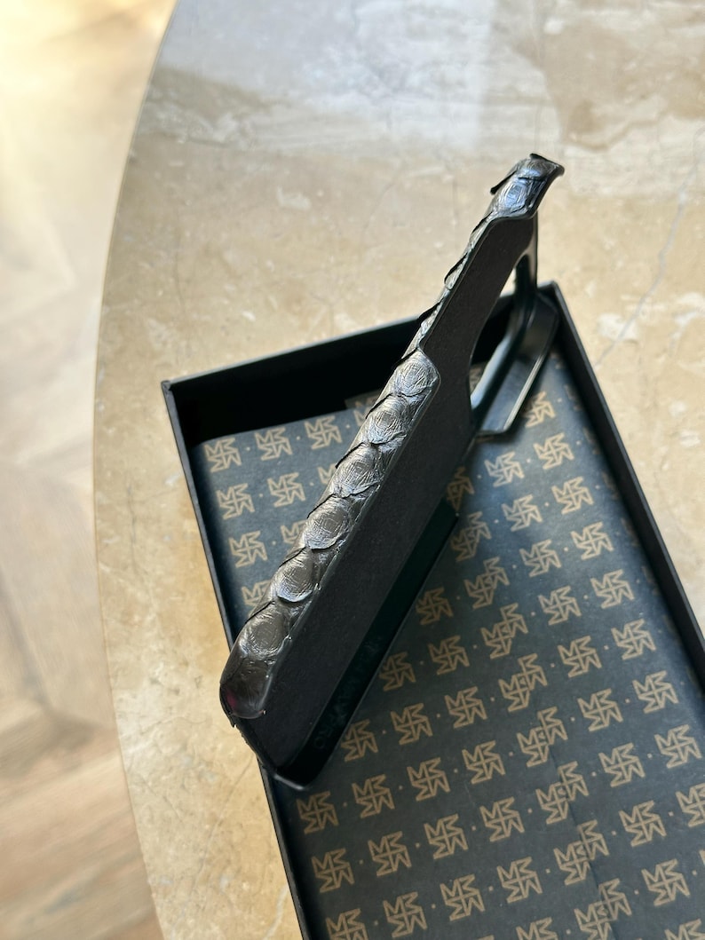 May include: A dark gray, textured letter opener with a decorative handle, resting inside a black box with a patterned interior. The handle has a scale-like design. The box's interior features a repeating pattern.