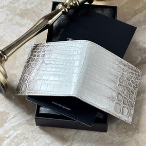Könnte beinhalten: Ein Faltgeldbörse mit einer weißen und grauen Krokodilleder-Textur. Die Geldbörse liegt auf einer schwarzen Schachtel, die eine Karte mit dem in Weiß gedruckten Text www.mhiraje.com enthält. Die Geldbörse hat eine glänzende Oberfläche.