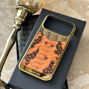 May include: A gold-colored phone case with an orange and brown snakeskin pattern. The case has a gold frame and the text "24k Gold Python Edition". The case is on top of a black box.