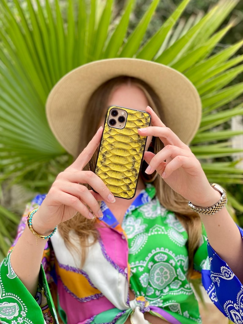 Pu&ograve; includere: Una custodia per telefono gialla con motivo a pelle di serpente e bordo nero. La custodia &egrave; tenuta davanti a una persona che indossa una camicia fantasia colorata e un cappello di paglia. Foglie di palma verdi sullo sfondo.