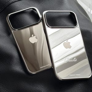 Puede incluir: Dos fundas plateadas para tel&eacute;fono con superficie reflectante. Cada funda tiene un marco rectangular y un logotipo de Apple. Una funda est&aacute; grabada con el nombre "Charlotte" y el texto "Platinum Edition". Las fundas se muestran sobre una superficie de cuero negro.