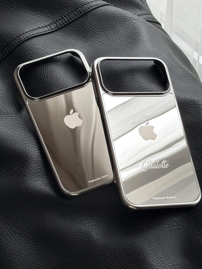 Puede incluir: Dos fundas plateadas para tel&eacute;fono con superficie reflectante. Cada funda tiene un marco rectangular y un logotipo de Apple. Una funda est&aacute; grabada con el nombre "Charlotte" y el texto "Platinum Edition". Las fundas se muestran sobre una superficie de cuero negro.