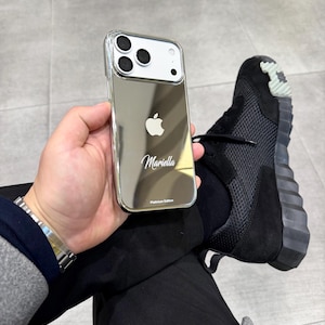 Puede incluir: Funda plateada para iPhone con acabado de espejo, con el logotipo de Apple y el nombre "Mariella". La funda est&aacute; etiquetada como "Platinum Edition". La imagen tambi&eacute;n muestra una zapatilla deportiva negra.