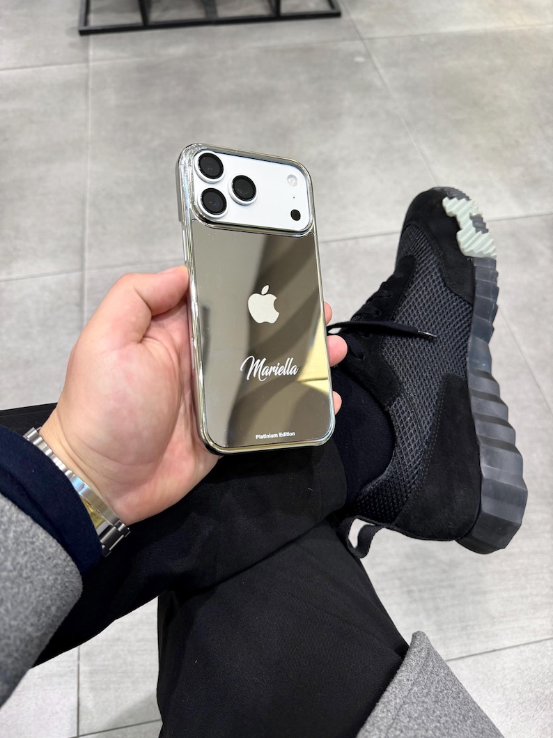 Puede incluir: Funda plateada para iPhone con acabado de espejo, con el logotipo de Apple y el nombre "Mariella". La funda est&aacute; etiquetada como "Platinum Edition". La imagen tambi&eacute;n muestra una zapatilla deportiva negra.