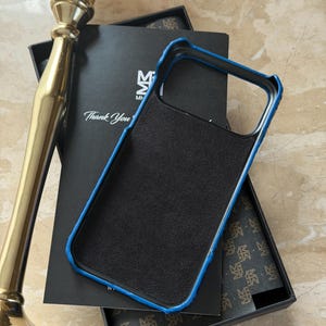 May include: Black phone case with a blue trim, resting on a black box. The case has a textured surface. The box has the text "Thank You" and a logo. A gold-colored decorative object is in the background.