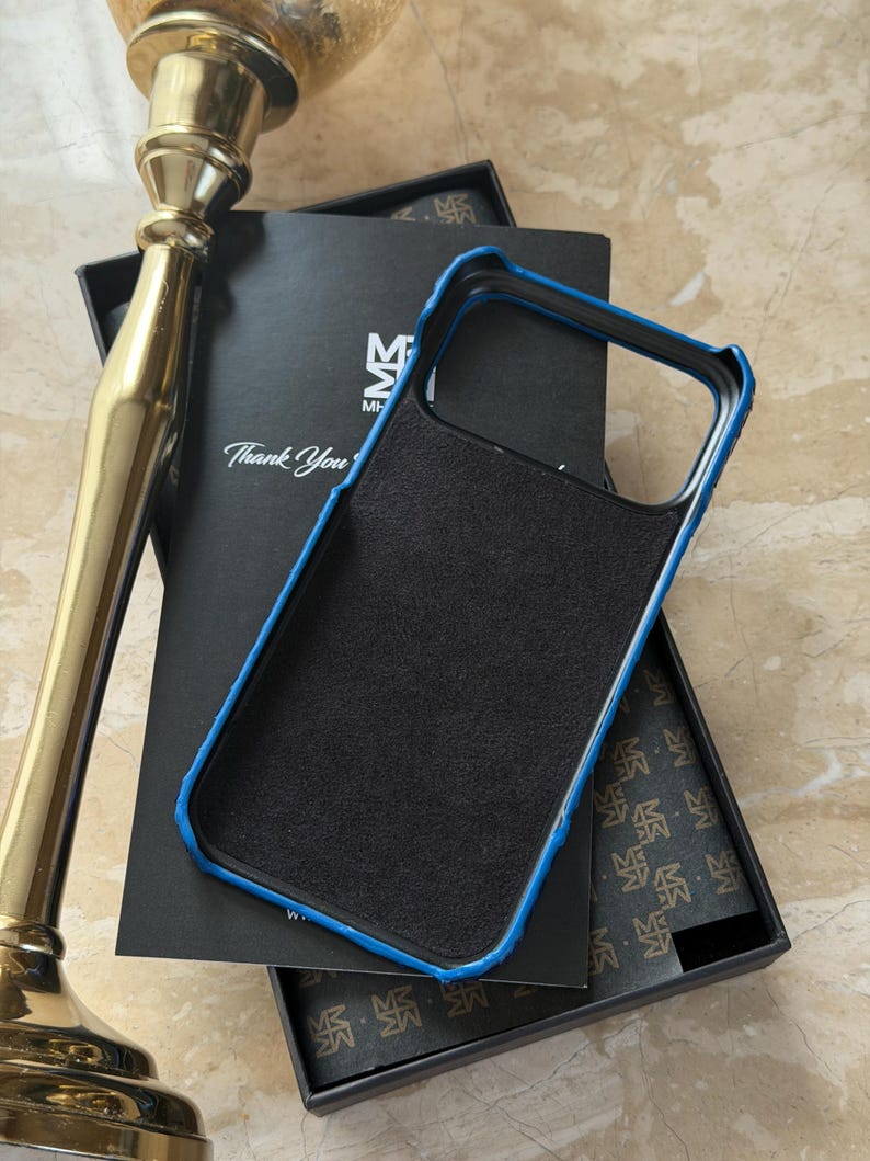 May include: Black phone case with a blue trim, resting on a black box. The case has a textured surface. The box has the text "Thank You" and a logo. A gold-colored decorative object is in the background.