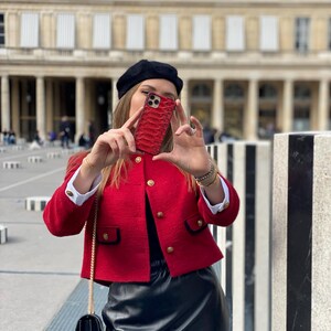 May include: A red textured phone case with a snake skin pattern. The case is being held up in front of a person. The person is wearing a red jacket with gold buttons, a black beret, and a black skirt.