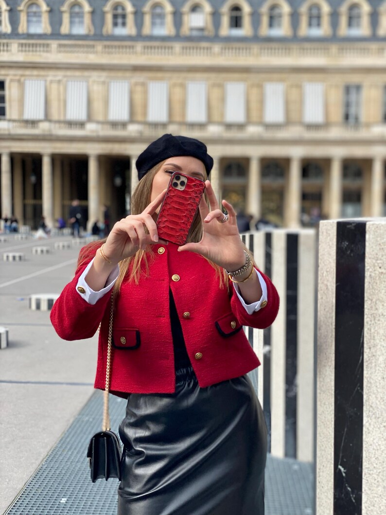 May include: A red textured phone case with a snake skin pattern. The case is being held up in front of a person. The person is wearing a red jacket with gold buttons, a black beret, and a black skirt.
