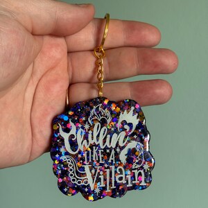 May include: A purple, black, and orange glitter keychain with a white outline of the text "Chillin' Like a Villain".