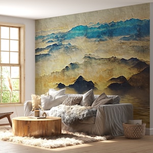 May include: A living room interior design featuring a large wall mural depicting a watercolour-style landscape of mountains and clouds in shades of blue, yellow, and brown. The mural is above a grey sofa with throw pillows and a faux fur throw. A wooden coffee table sits in front of the sofa.