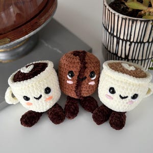 May include: Three crocheted coffee-themed plushies: two coffee cups and a coffee bean. The cups are white with brown coffee and have smiling faces. The bean is brown with a smiling face. All have brown legs.