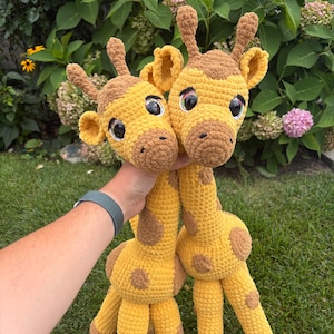 Puede incluir: Dos jirafas de crochet hechas a mano. Las jirafas son amarillas con manchas y detalles marrones. Tienen ojos grandes y negros y orejas pequeñas. Las jirafas se sostienen frente a un fondo verde.
