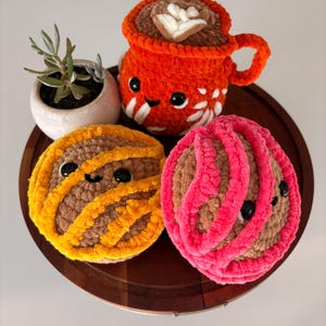 May include: A collection of handmade crochet items, including an orange coffee cup, two round pastries, and a small potted succulent. The coffee cup has a smiling face and white accents. The pastries are brown with yellow and pink accents.