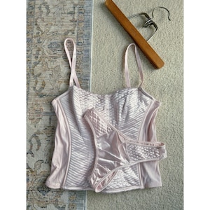 May include: A light pink lingerie set, including a bustier top and matching thong. The bustier features a quilted pattern on the front and adjustable straps. The thong has a similar quilted design. A wooden hanger rests above the set.
